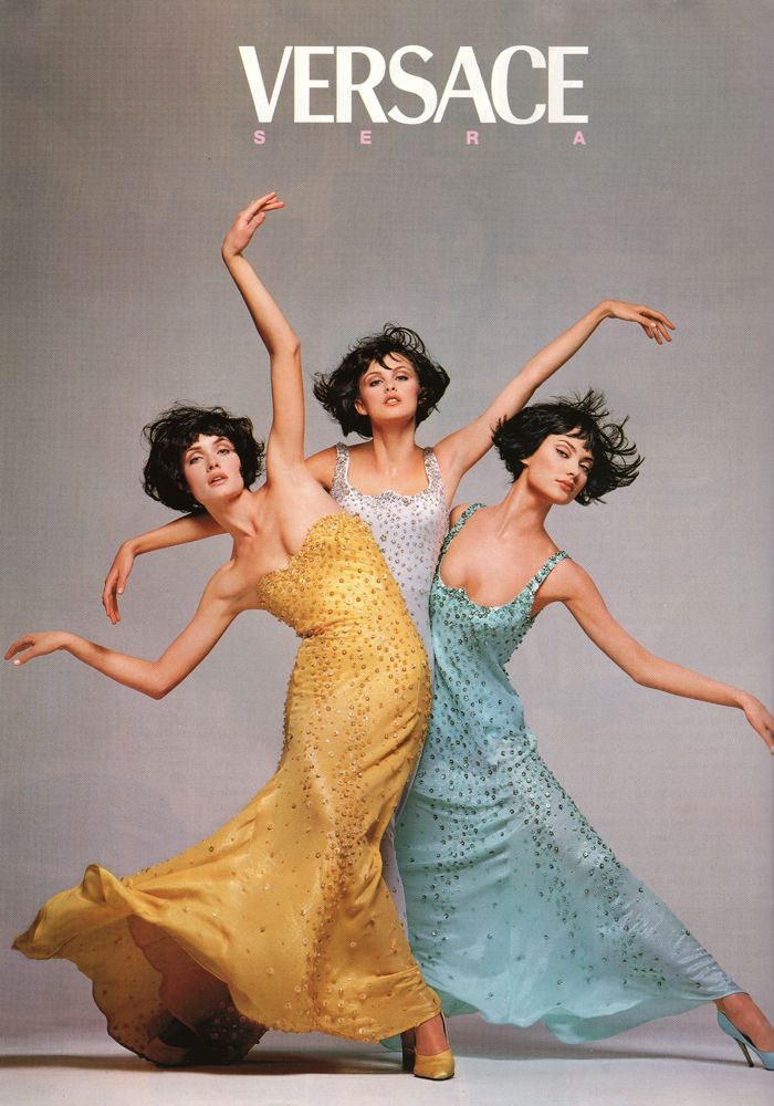 three fashion models pose in a studio for a versace ad. the models are wearing long flowing dresses that appear to be beaded, with one in yellow, one in aqua blue, and one in silver. the models are holding dramatic, elegant poses. the models are all looking at the camera with a serious expression.