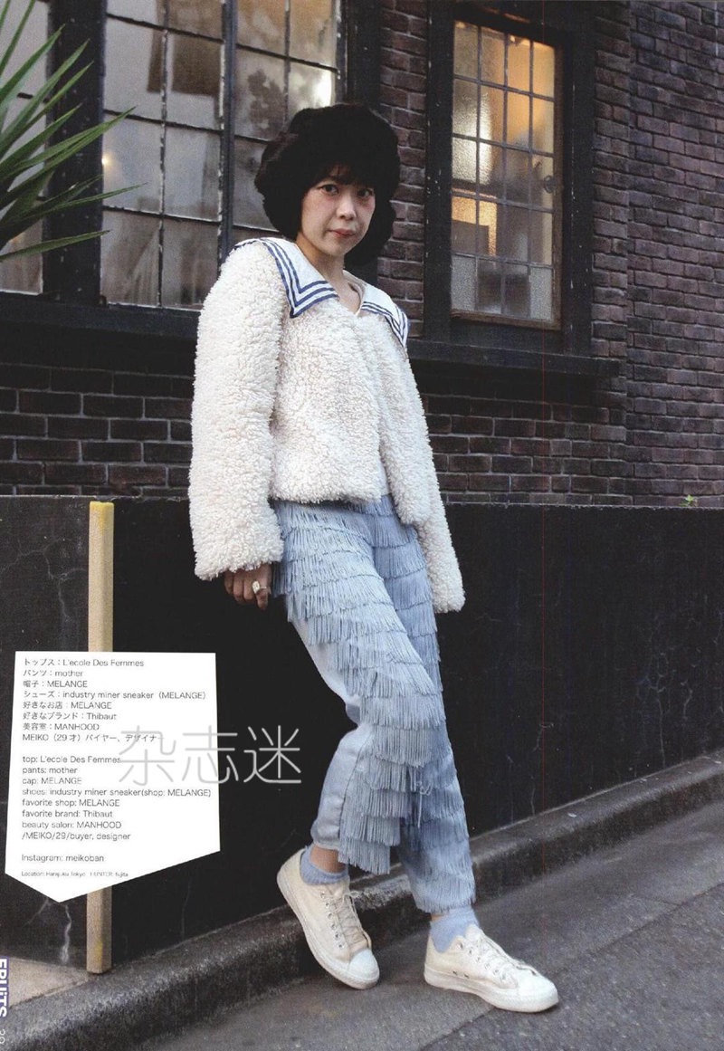 a woman in a white fleece jacket, light blue jeans with fringe detail, white sneakers, and a black hat poses for a street fashion photograph. she is leaning against a pole in front of a brick building with a window.  the photo appears to be from a japanese fashion magazine.