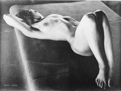 the image is a black and white photograph of a nude female model reclining on a darkcolored upholstered surface. the model is posed gracefully, with her arms extended behind her head. the lighting in the photograph is dramatic, emphasizing the curves and contours of her body through strong shadows and highlights. the photograph appears to be a vintage print, possibly from the mid20th century, showing a style consistent with art photography of that period. the overall mood is both sensual and artistic, focusing on the beauty of the human form.  the texture of the surface beneath the model is visible, contrasting with the smooth skin of the model.