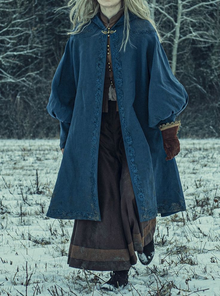 a woman in a blue and brown medievalstyle cloak walks through a snowy forest. the woman has long blonde hair and is wearing a long, blue cloak with embroidery. she is also wearing a brown skirt. the woman is walking in a forest, with snow on the ground. the forest is dark and mysterious, and there are trees in the background.