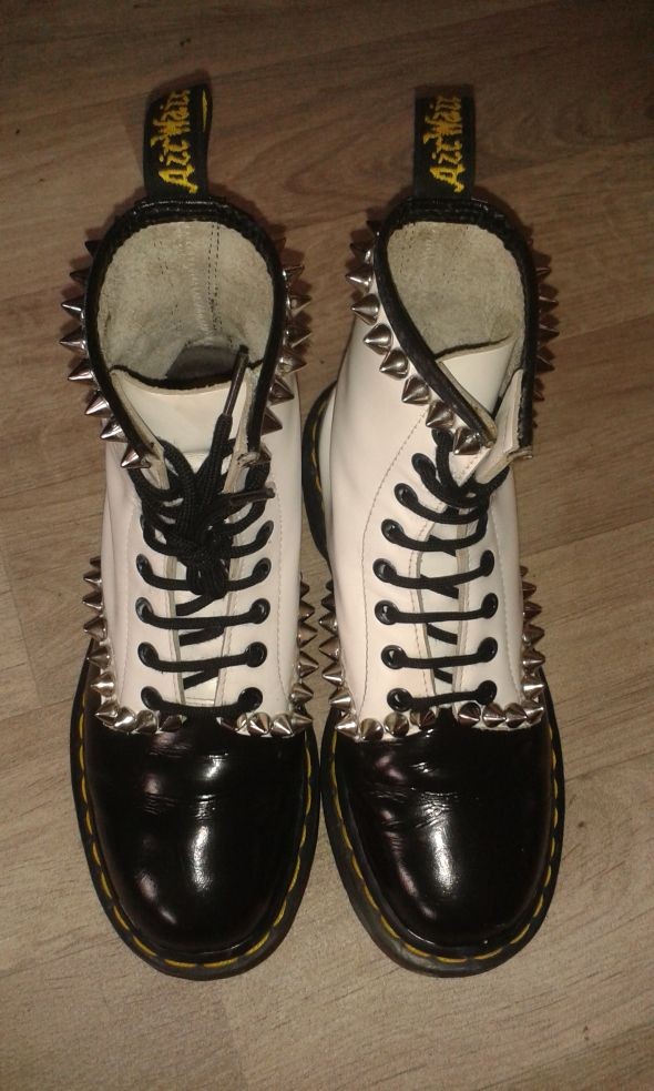 a pair of white and black dr. martens boots with studs all around. the boots are laceup and have a black leather toe cap.  the boots are sitting on a wooden surface.