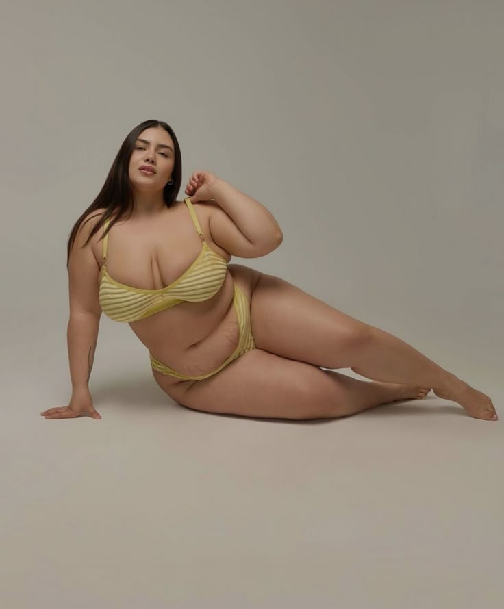 a plussize woman is lying down in a studio wearing yellow striped lingerie. she has dark hair and is posed confidently. the image promotes body positivity.