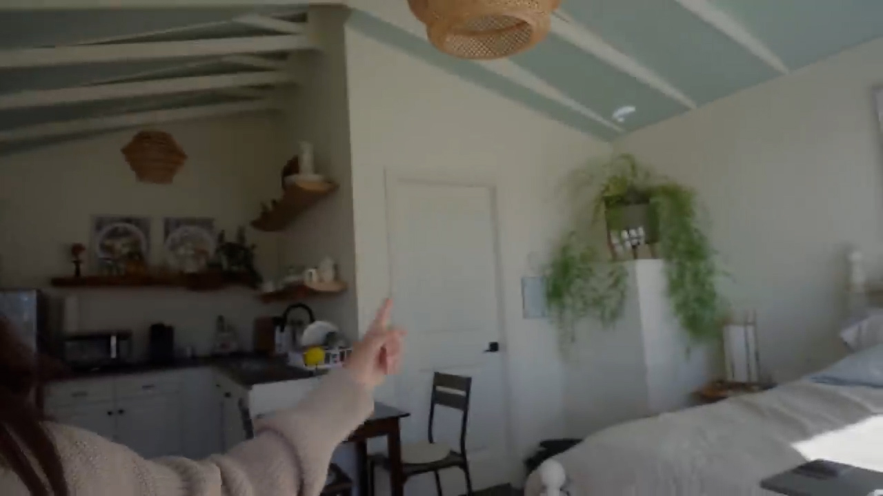 a woman stands in a small, bright room with a vaulted ceiling painted a light bluegreen.  she is pointing to her right, showing off the space which appears to be a combined bedroom and kitchenette. to the left is a small kitchen area with white cabinets, open shelving, and a countertop. to the right is a bed with white linens and a tall plant in the corner. the room is decorated in a minimalist style with neutral colors and natural materials.