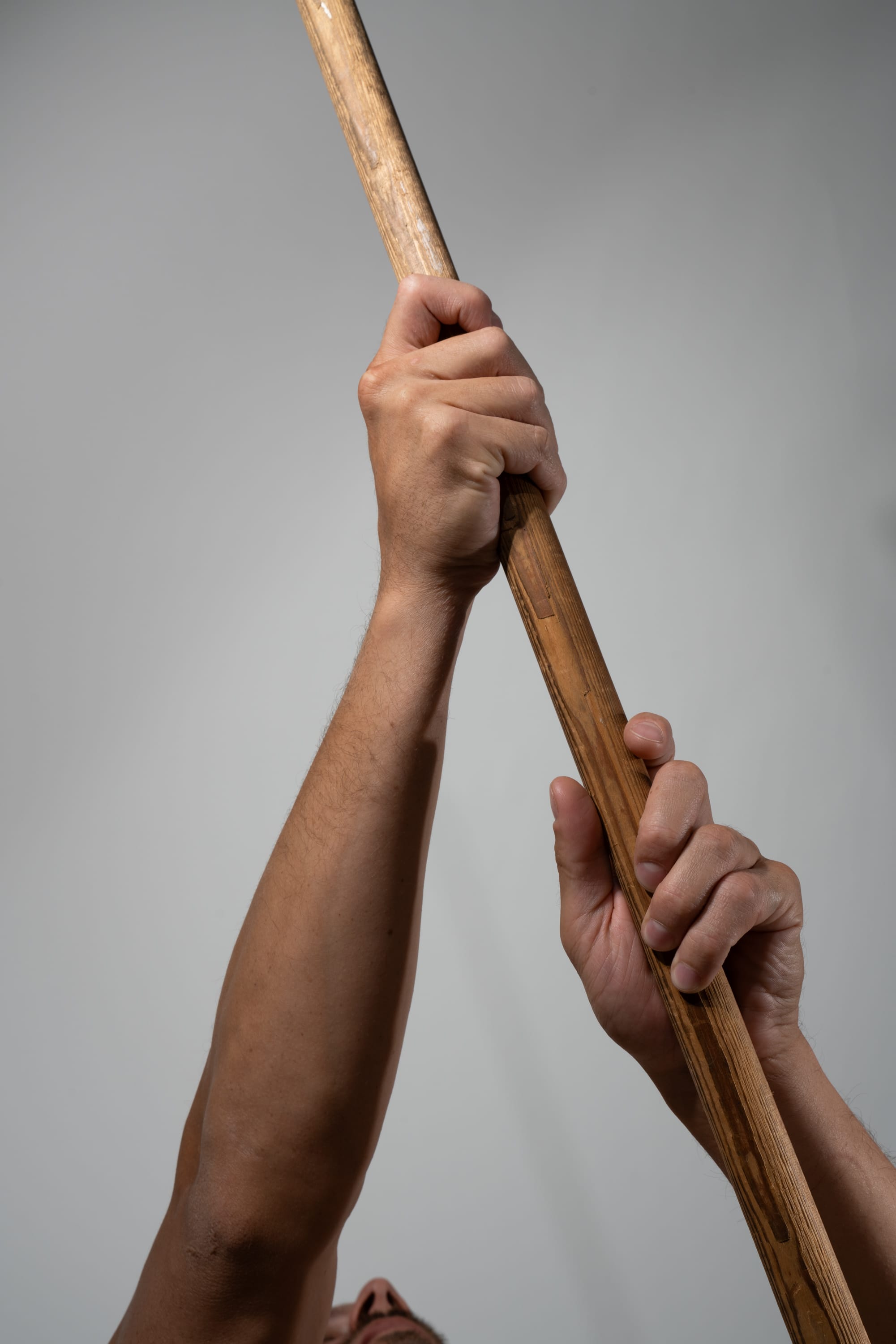 a closeup, lowangle view of a man's arms as he powerfully grips a long wooden pole, lifting it towards the ceiling. the focus is on the strength and tension in his hands and forearms.