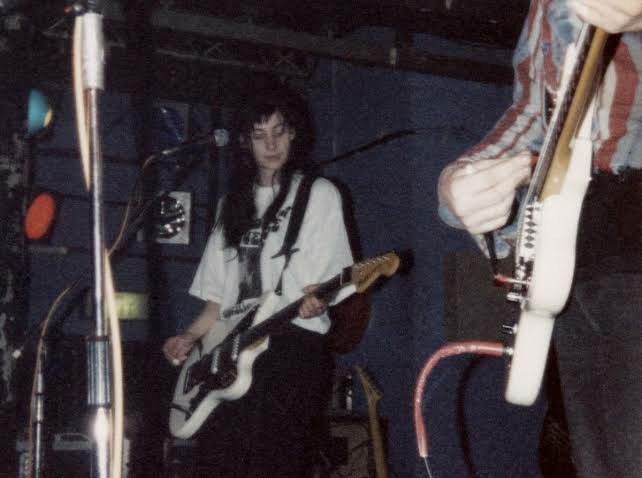 a blurry, candid photo from the 90s showing a female guitarist on stage, performing a live rock concert. she has long dark hair and is wearing a white tshirt.  she's playing a white electric guitar. another person, partially visible, is also playing a guitar, wearing a striped shirt. the background is dark, with some lighting on the stage. the overall atmosphere is reminiscent of the grunge or alternative rock scene of that era. the image has a grainy, vintage quality.