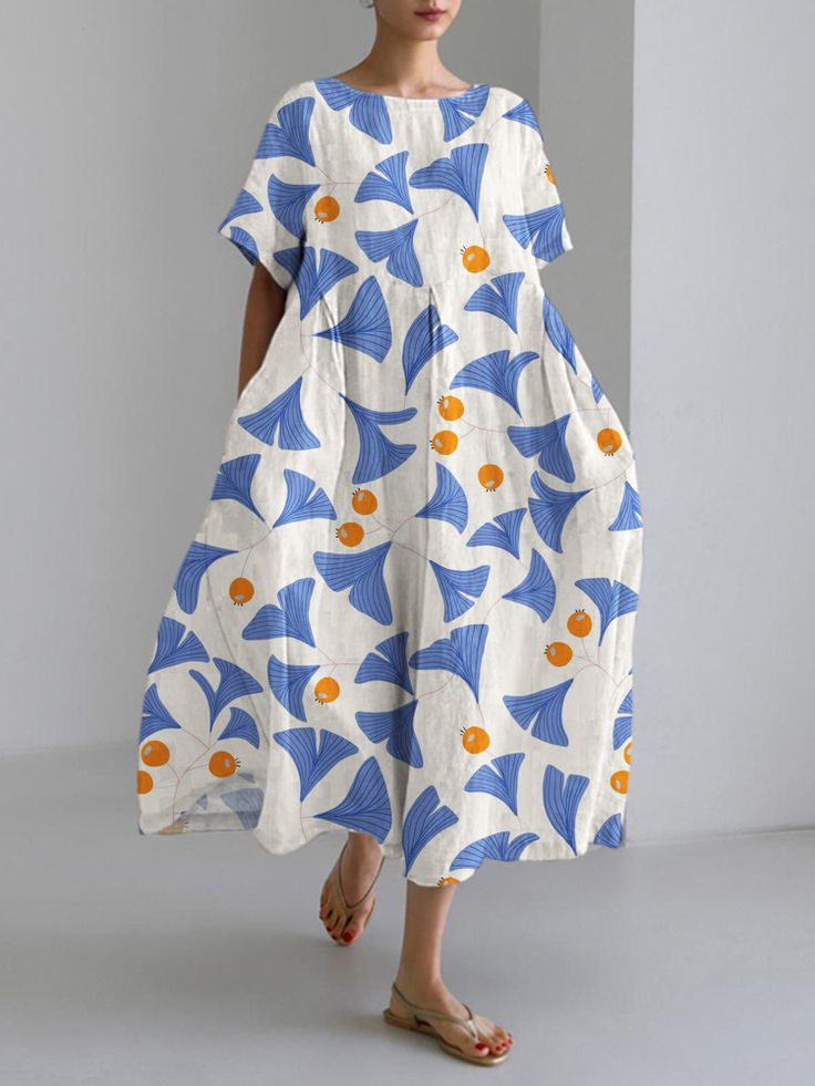 a woman wearing a white and blue floral dress is standing and looking down. the dress has a ginkgo leaf pattern and an orange color detail.  she is wearing flat sandals.