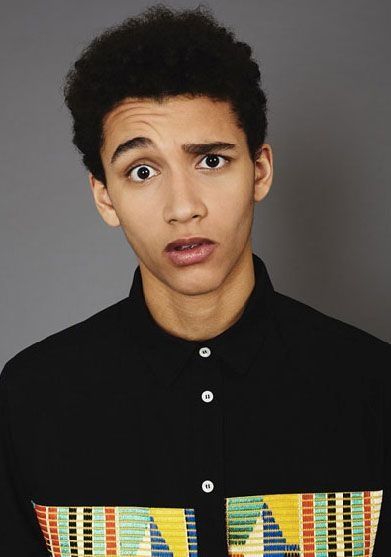 a young black man with short black hair is looking directly at the camera with a curious expression. he is wearing a black collared shirt with colorful buttons and a patterned bottom, and he has a serious look on his face.  the photo was taken in a studio setting against a gray background.