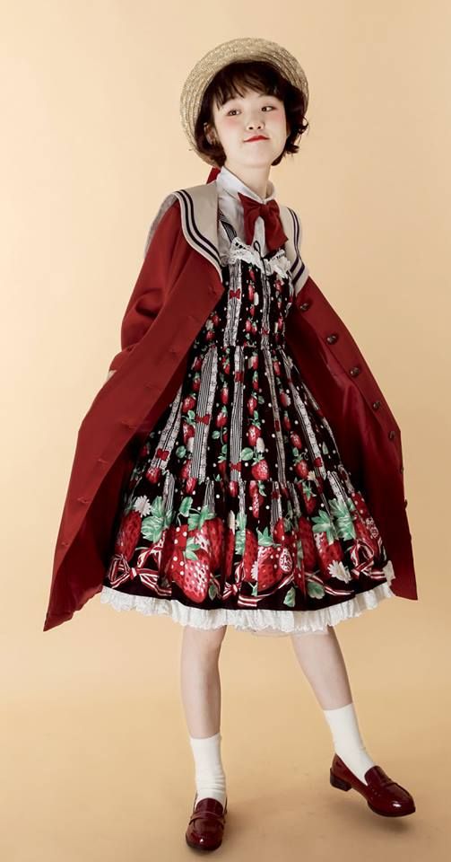 a young girl in a red and white dress with strawberries on it is standing in front of a beige background. she is wearing a straw hat and red shoes. she has a cute and stylish look.