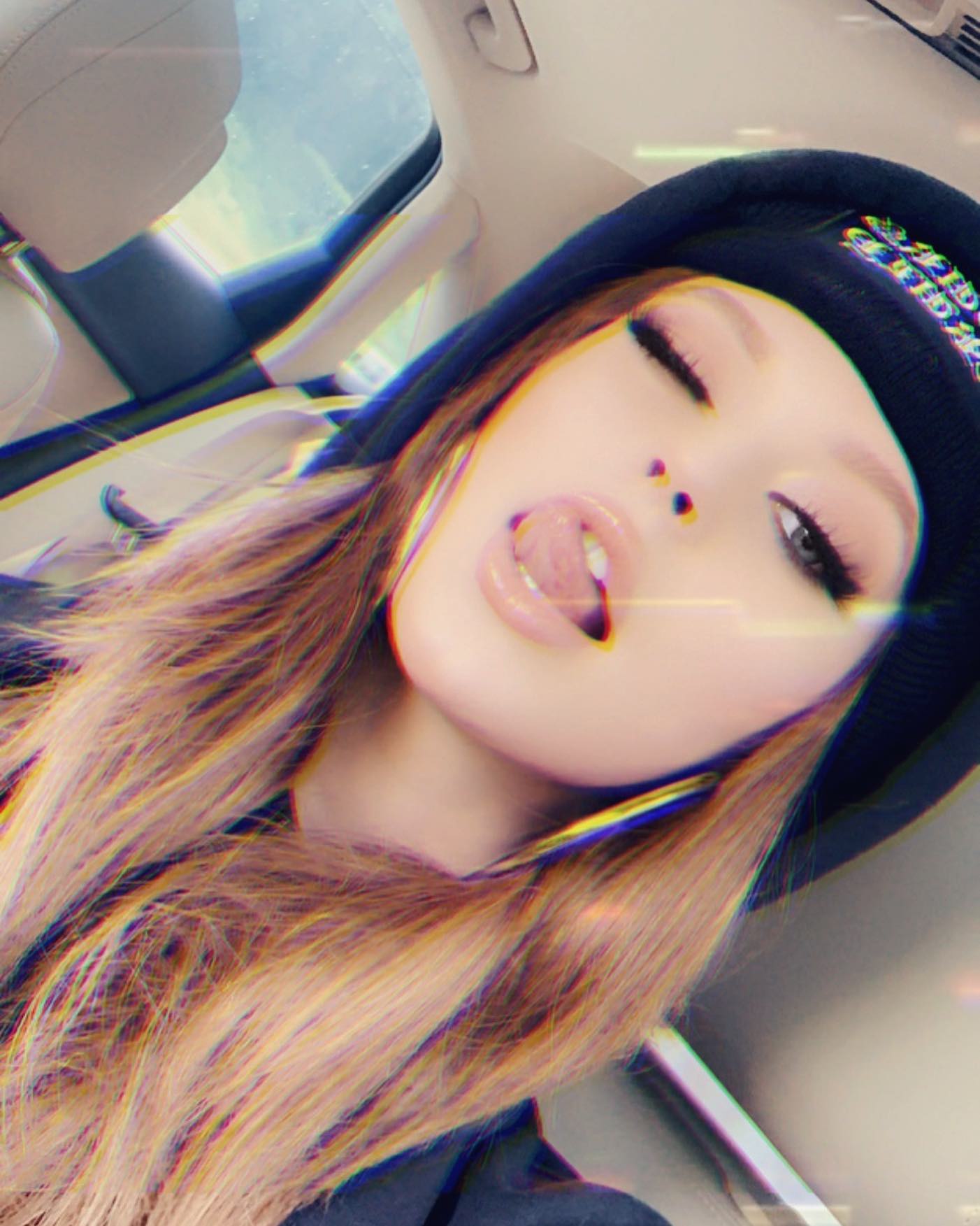 a selfie of a woman with her tongue out wearing a beanie, hoop earrings, and makeup in a car.