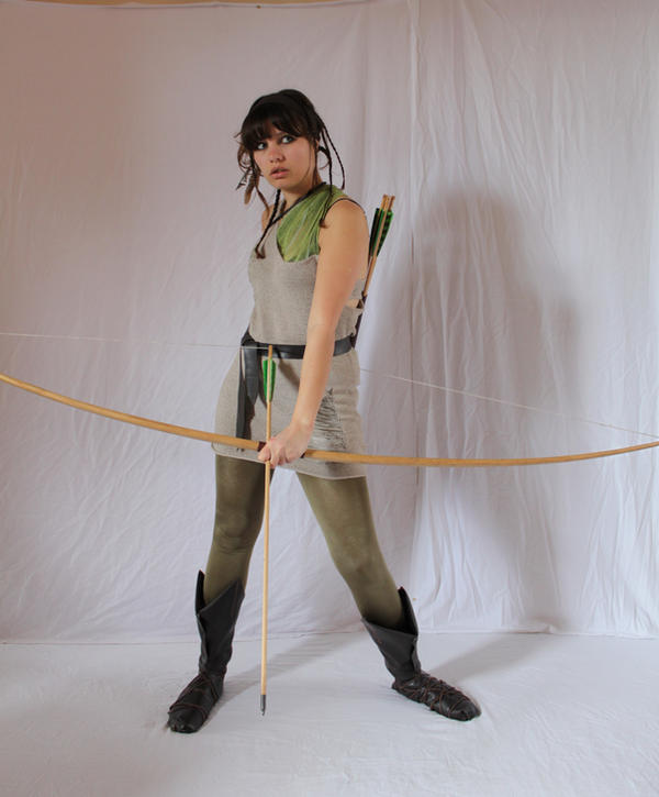 a fullbody studio shot of a young woman dressed as an archer. she is wearing a light beige tunic, green leggings, and brown leather boots.  she holds a longbow and arrow, with a quiver of arrows on her back. she has dark hair and is looking directly at the camera. the background is a simple white fabric backdrop. she is in a ready stance.