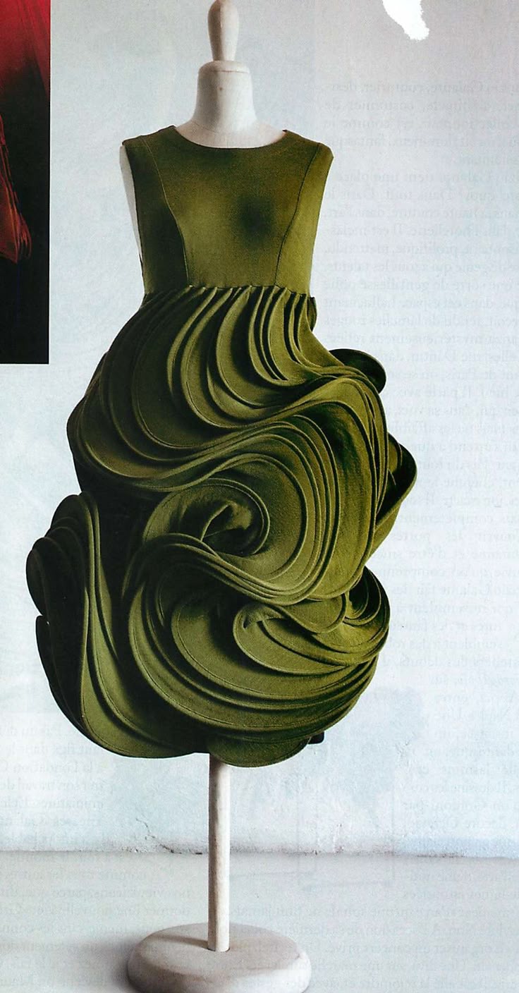 an olive green dress is displayed on a white mannequin. the top portion of the dress is fitted, while the skirt is a complex arrangement of swirling, sculpted fabric. the dress is displayed in front of a white wall.