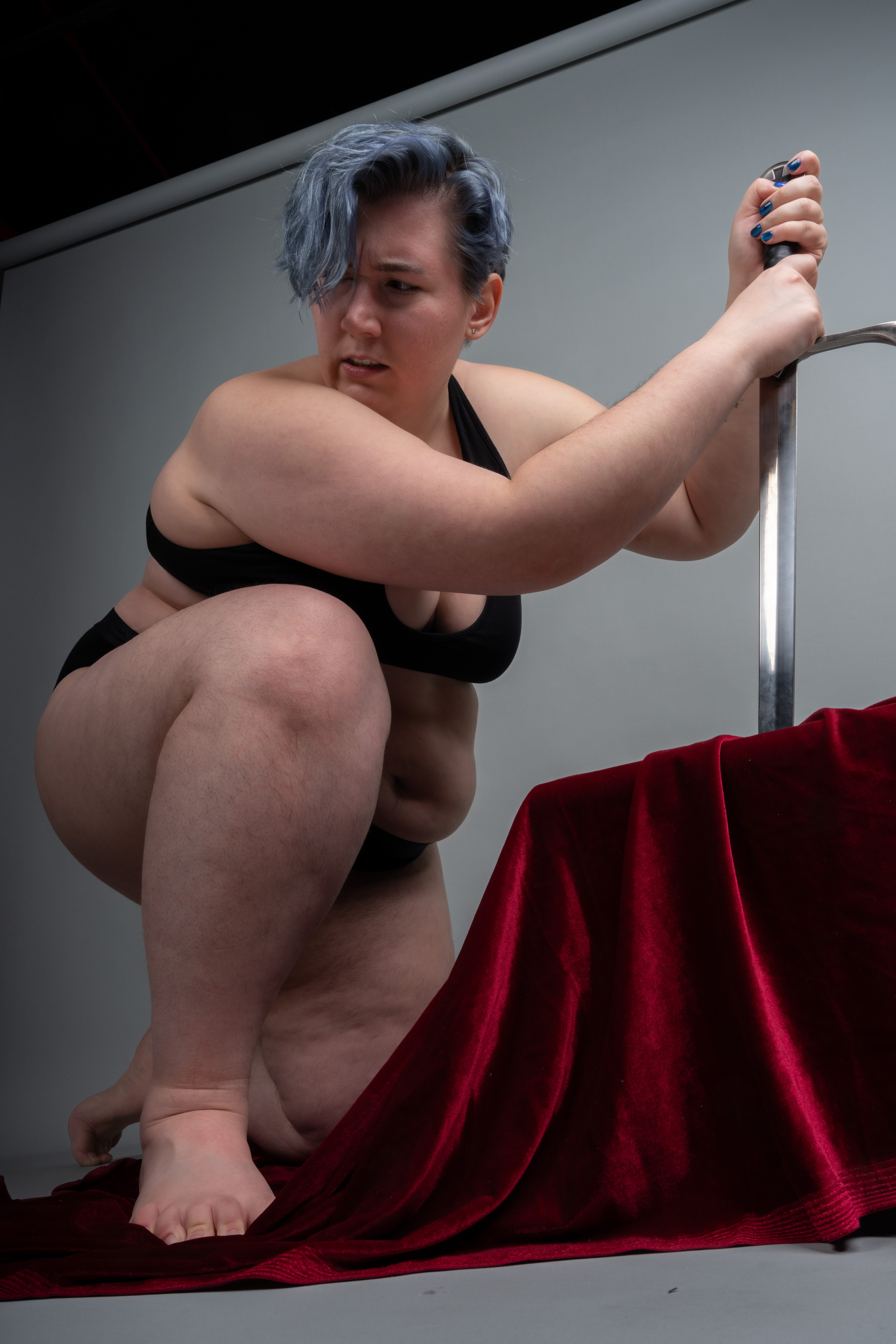 a plus size woman with short blue hair wearing a black bra and underwear is kneeling indoors while holding a sword and looking to the side in fear. a red velvet cloth is draped in front of her.