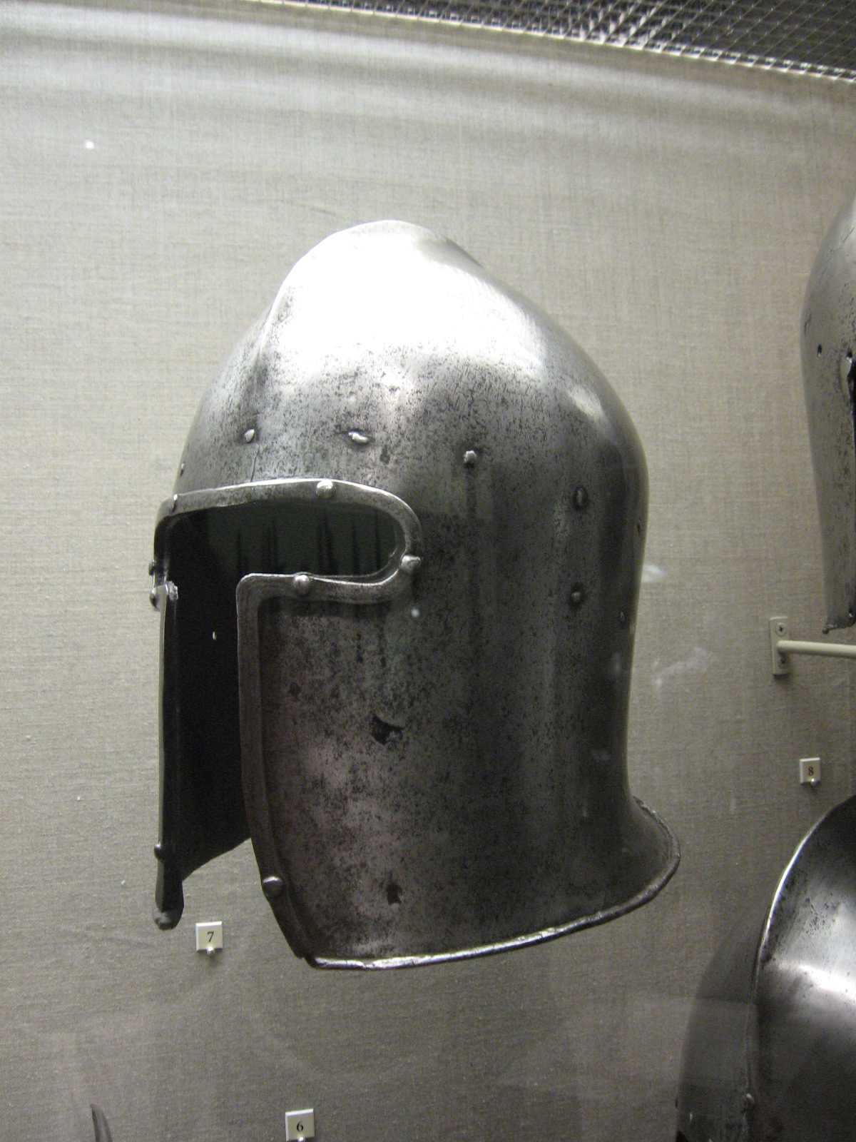 the image shows a closeup view of a medieval helmet displayed in a museum. the helmet is made of polished steel and has a simple, functional design. it features a narrow visor with a horizontal slit for vision and a slightly pointed crown. the helmet is mounted on a plain gray wall, and the lighting is artificial, casting some shadows on the object. the overall impression is one of history and craftsmanship.