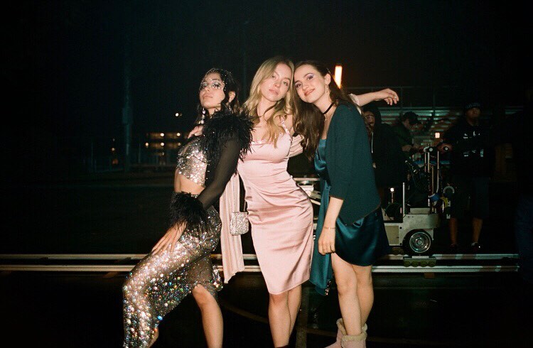 three young women are standing close together outside at night, possibly on a film set. they're dressed in glamorous party attire. one girl is wearing a sparkly silver dress, another a pale pink satin dress, and the third a teal silk dress.  they're smiling and posing for a photo. behind them, a film crew and equipment are visible in the blurry background, suggesting that this is a behindthescenes shot. the overall atmosphere is dark and glamorous.