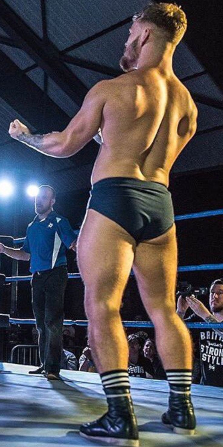 the image shows a muscular male wrestler from behind, standing in a wrestling ring. he is wearing black wrestling trunks and black and white socks with boxingstyle boots. his back and legs are prominently displayed, showcasing his athletic physique.  a referee is visible in the background, as is a portion of the audience. the overall setting suggests a wrestling match or event is taking place. the lighting highlights the wrestler's musculature.