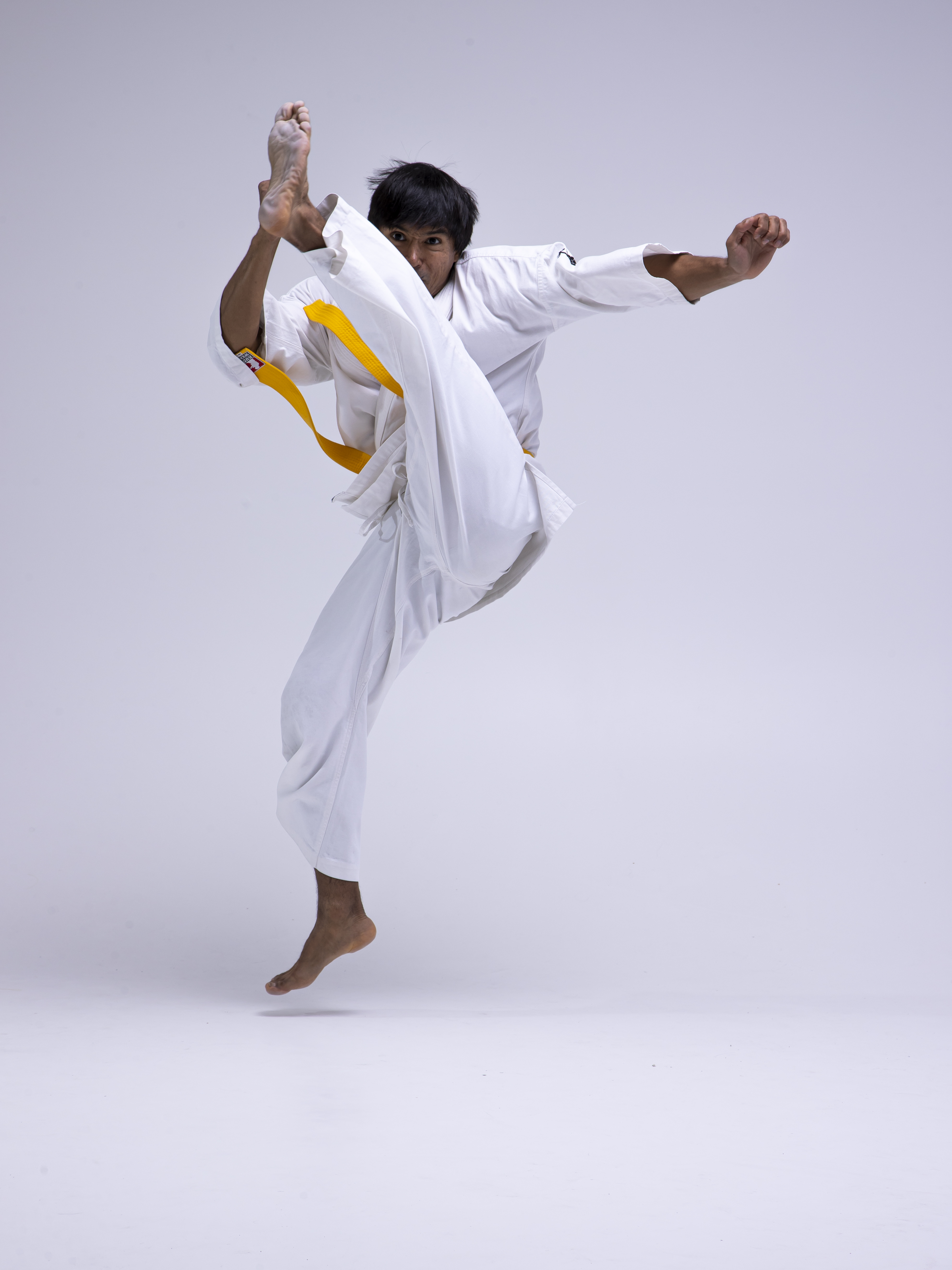 a man wearing a white karate uniform with a yellow belt is captured midair performing a high kick. he is barefoot, with one leg extended upwards and the other providing support. his hands are positioned in a defensive manner, suggesting a dynamic action shot in a studio setting.