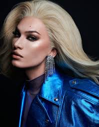 the image is a closeup portrait of a woman with long, platinum blonde hair styled in a voluminous, somewhat disheveled way.  she's wearing a vibrant blue, shiny leather jacket and dangling crystal earrings. her makeup is emphasized, including a strong eye look.  the lighting is dramatic, with a focus on her face and hair against a dark background. the overall aesthetic is glamorous and fashionforward. the woman appears to be a model.