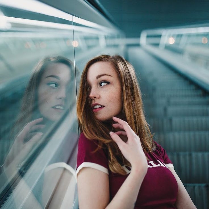 a young woman with long, red hair is captured in a candid moment, leaning against the mirrored wall of an escalator. she's stylishly attired in a maroon top with white trim, her gaze drawn towards her reflection. the photo's composition cleverly uses the escalator's receding lines and the play of light and reflections to create a sense of depth and movement. her relaxed posture and enigmatic expression invite the viewer to wonder about her thoughts and journey.