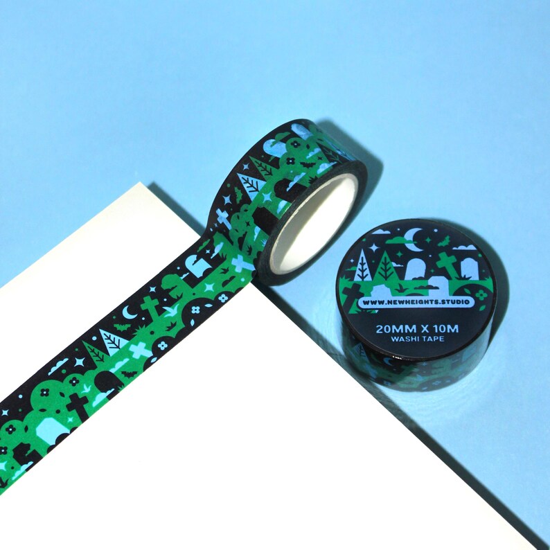a roll of washi tape featuring a graveyard scene with black trees, crosses, moons, and stars on a green background. the tape is 20mm wide and 10m long. the tape is made by new heights studio and is displayed on a white surface against a light blue background.