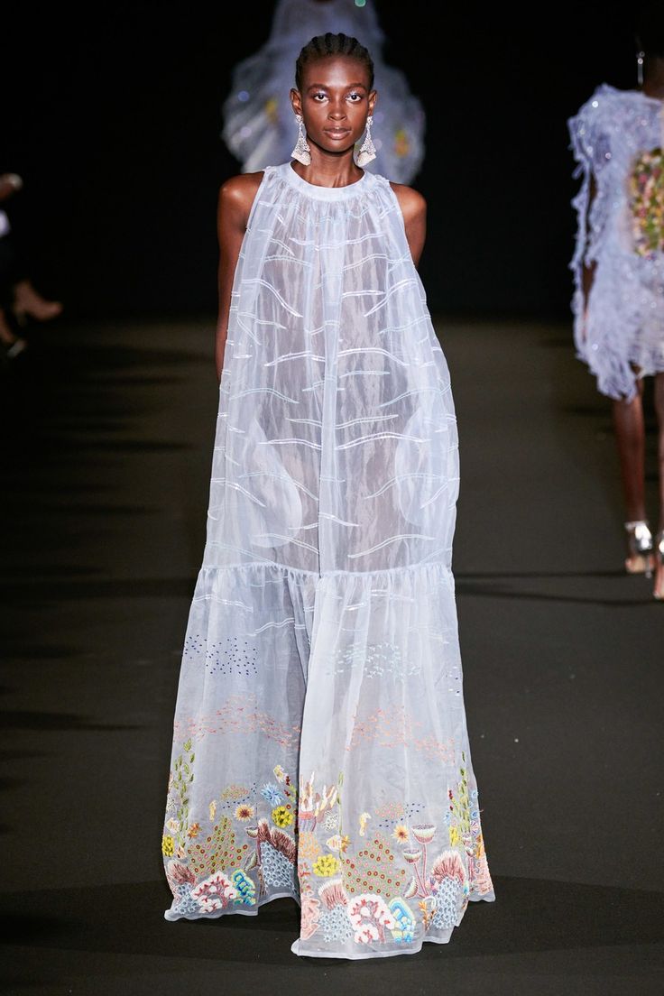 a model walks the runway in a white, sheer, long dress with delicate floral embroidery. the dress is very flowing and elegant. the model is walking confidently and has a beautiful, ethereal look. the overall image is one of high fashion and sophistication.