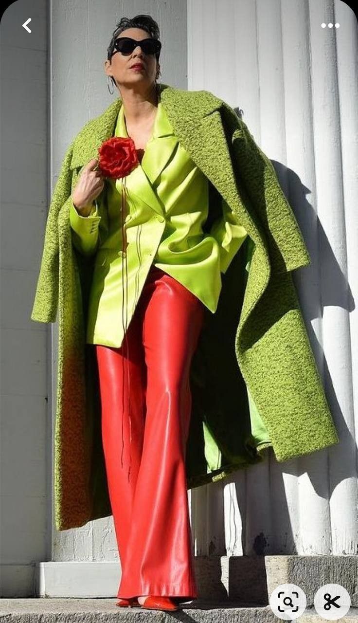 a woman is standing on a step in front of a building. she is wearing a green coat over a green blazer and red pants. she is also wearing sunglasses and holding a red flower.