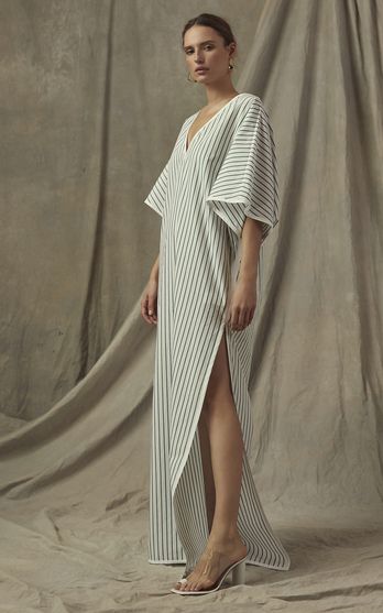 a full shot of a fashion model wearing a long, striped maxi dress. the dress is white with thin, dark stripes and has a relaxed fit. it features a vneckline and a side slit. the model is wearing clear, highheeled sandals and is posing against a beige fabric backdrop. the overall aesthetic is minimalist and chic.
