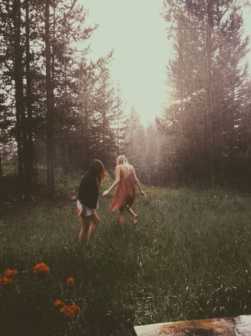 two women are walking through a grassy field in a forest on a rainy day. the sun is shining through the trees, creating a soft and blurred effect. there are orange flowers in the foreground. the women appear to be carefree and joyful.