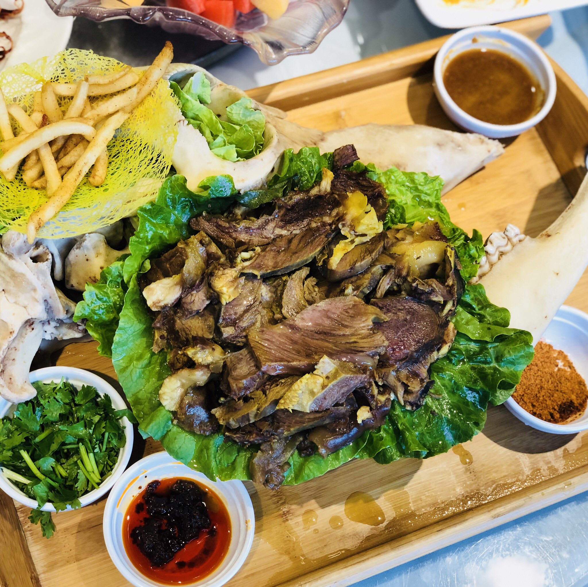 a closeup top view photo of a platter of lamb with the skull on the side. there are side dishes of lettuce, cilantro, dipping sauces, and french fries.
