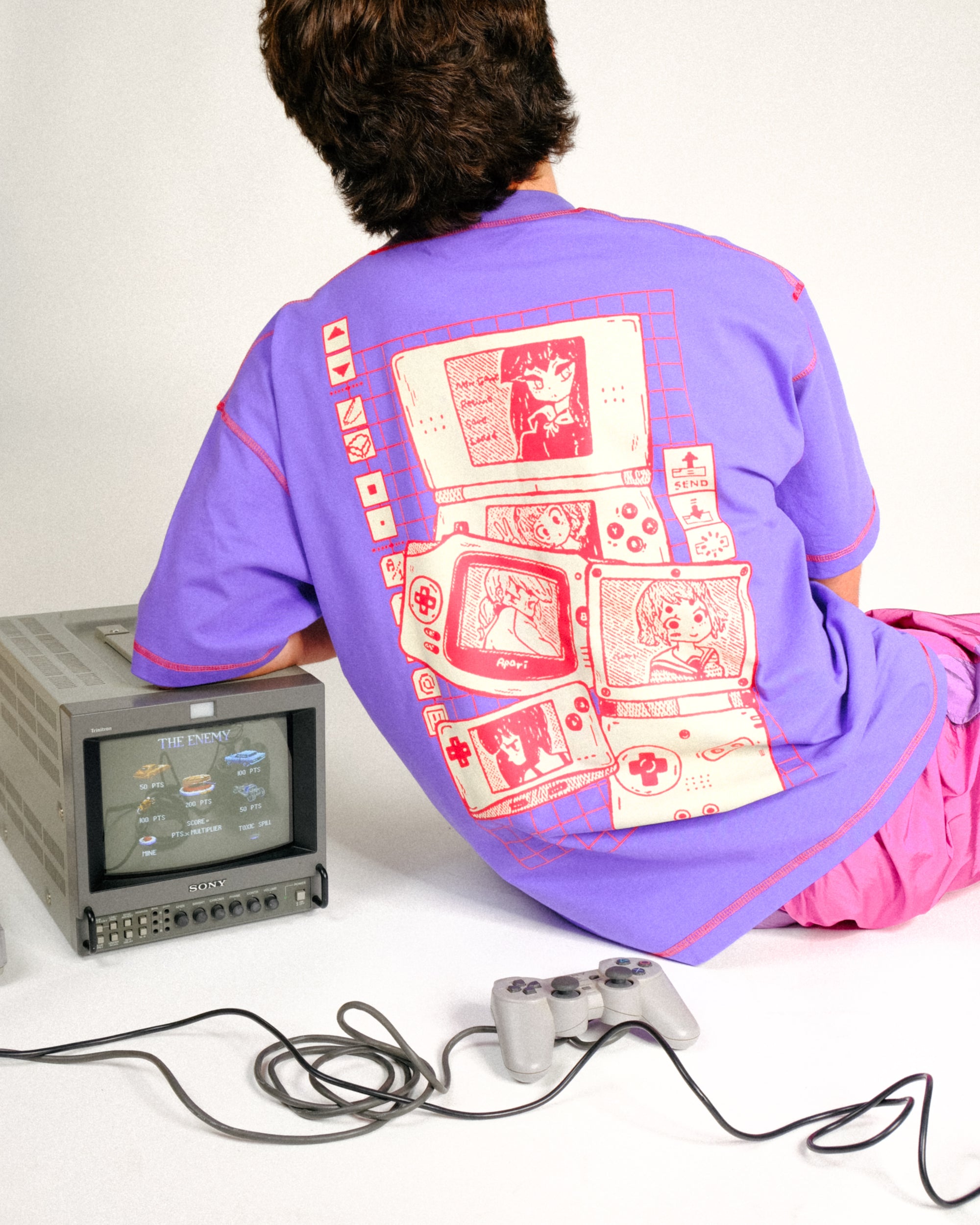 a person is sitting on the floor with their back to the camera, wearing a purple tshirt with a pink video game print, and pink pants. next to them is a gray sony monitor with a video game displayed on the screen, and a gray video game controller with a black cable. the cable is tangled on the floor. the background is plain white.