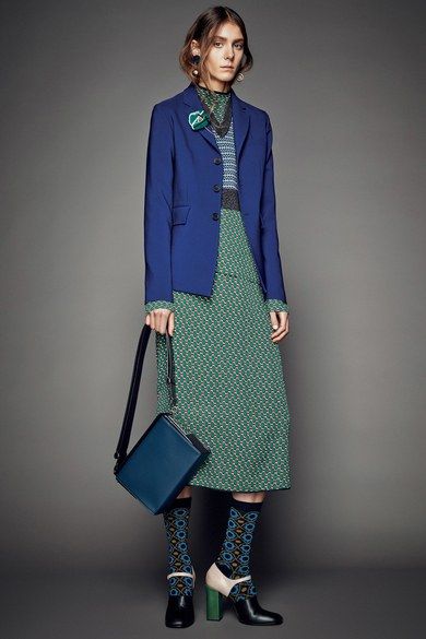 the image showcases a fashion model dressed in a stylish autumnal outfit. she is wearing a cobalt blue blazer over a green geometric patterned midi skirt and a patterned sweater.  the model is accessorized with patterned socks, highheeled shoes (one teal and one offwhite), and a dark blue crossbody bag. the overall style is sophisticated and elegant. the background is a neutral gray, keeping the focus on the model's attire.  the setting appears to be a professional photoshoot.