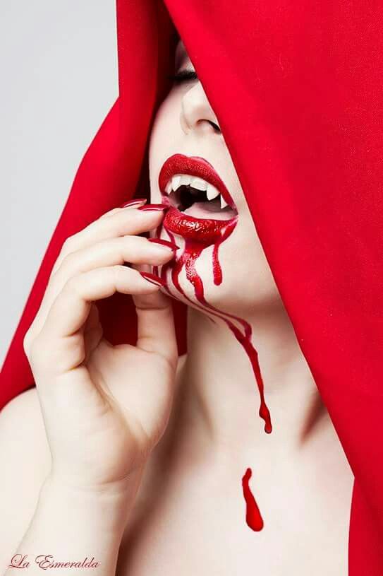the image shows a closeup portrait of a woman styled as a vampire.  she's wearing a vibrant red hooded cloak that partially obscures her face.  her makeup is dramatic, featuring deep red lipstick, visible fangs, and streaks of fake blood running down her chin and neck.  her expression is alluring and slightly sinister. the overall aesthetic is gothic and sensual, suggestive of a vampire theme.  the watermark "la esmeralda" is visible in the bottom left corner, suggesting the photographer's name or the model's name or alias.