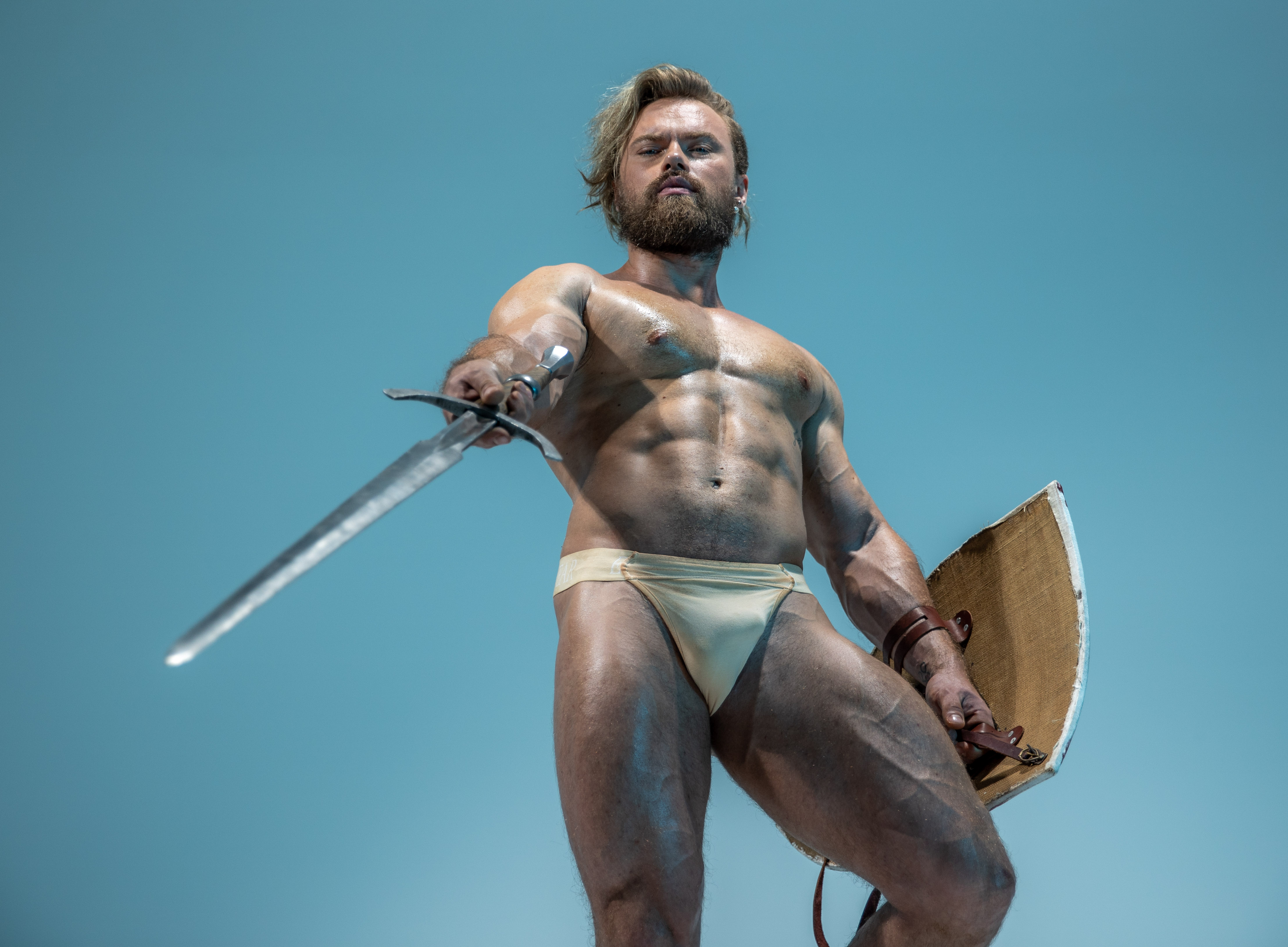 a muscular man with a beard and long blond hair stands in a warriorlike pose against a light blue background. he wears only a  light brown loincloth and holds a long, sharp sword pointed forward in his right hand. in his left hand, he holds a brown leatherbound shield.  the image is taken from a low angle, emphasizing his powerful physique and commanding presence. the studio lighting accentuates his muscles and creates a dramatic atmosphere.