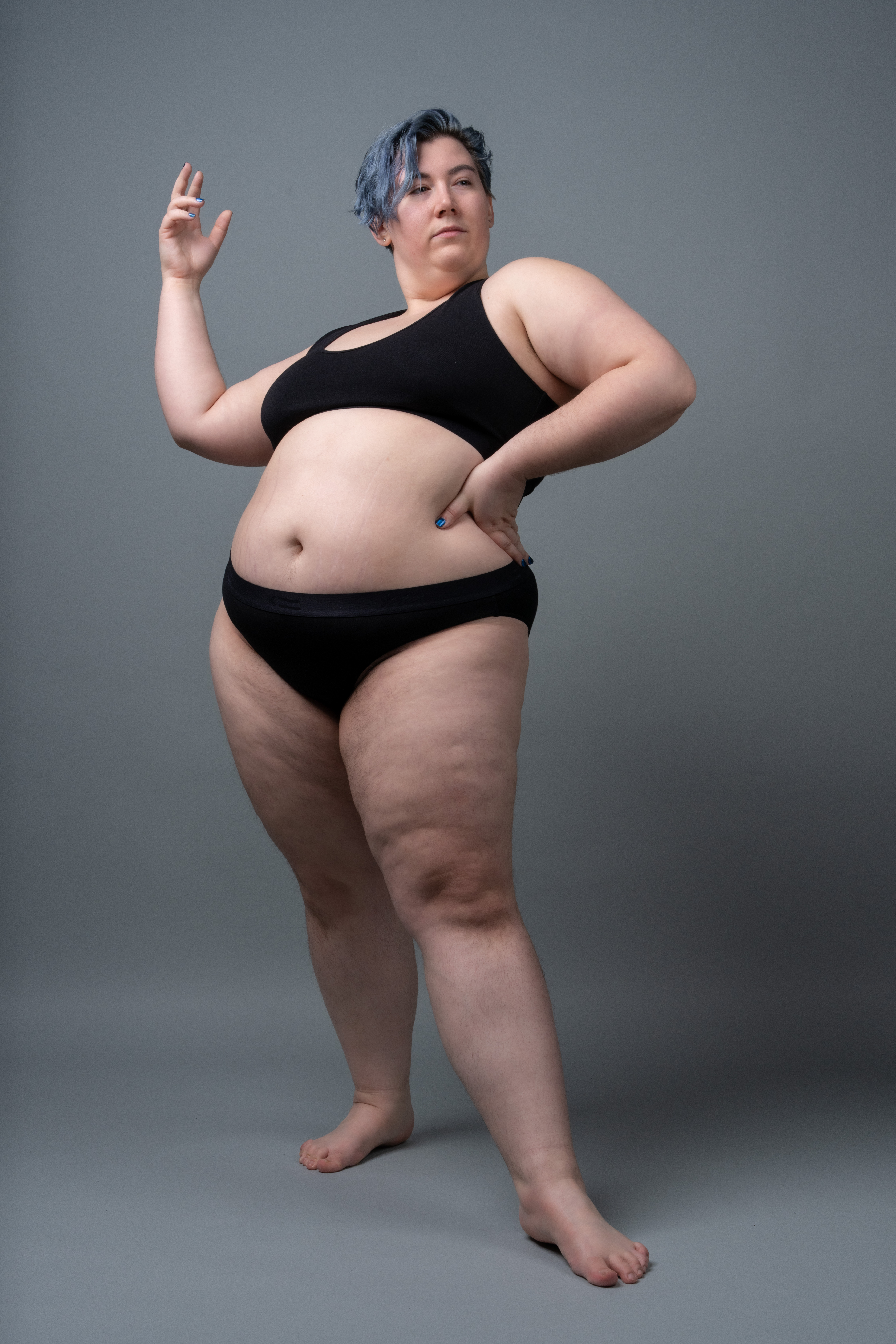 a fulllength studio shot features a plussize woman with blue hair, confidently posing against a gray background. she is wearing a black bra and black underwear, with one hand raised and the other resting on her hip. her posture exudes selfassurance and strength, promoting body positivity.