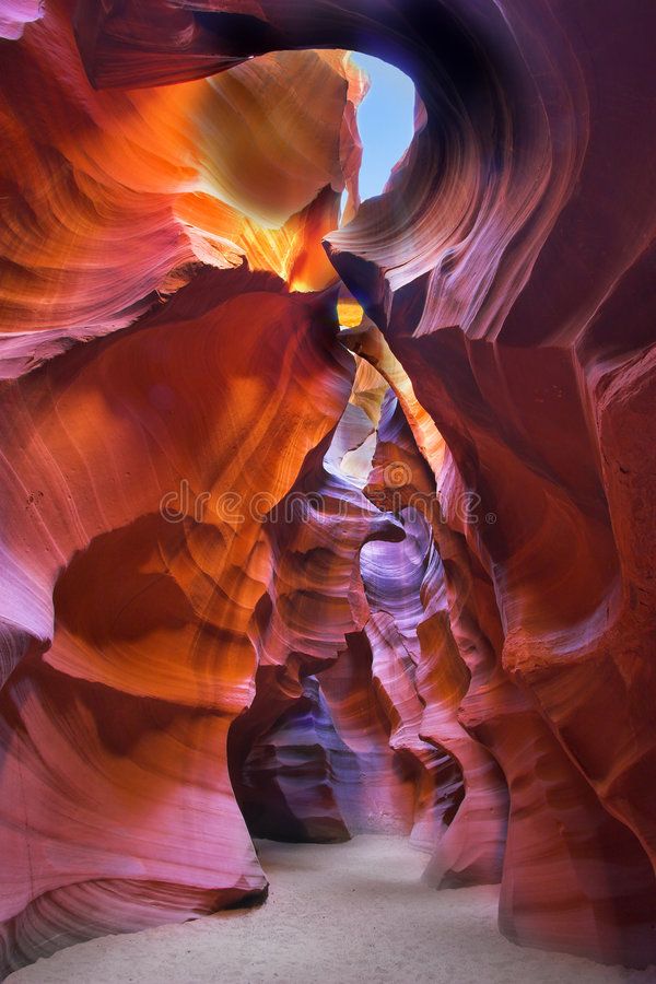 a view of a colorful slot canyon with red rock formations. the canyon is narrow and the walls are smooth, creating a beautiful, natural tunnel.  the sunlight shines into the canyon, illuminating the sandstone with a red, orange and purple hue.  the natural light makes the canyon appear otherworldly.  a thin layer of sand can be seen on the canyon floor.