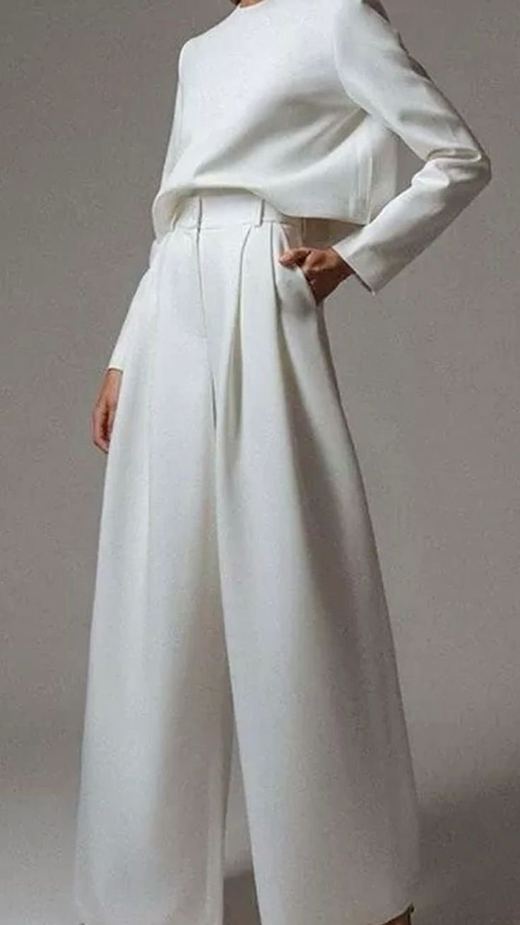 a woman is wearing a white dress with a long sleeve top and wideleg pants in a studio. she is posing with her hand in her pocket. the dress is simple and elegant. the model is wearing her hair pulled back and is wearing minimal makeup.