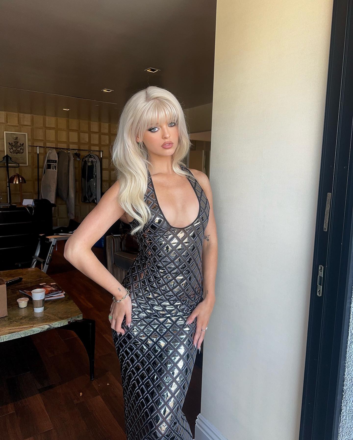 a fulllength shot of a woman with blonde hair and bangs, wearing a sequined silver halter dress. she is standing indoors, posing with one hand on her hip, and looking at the camera. she has jewelry on her wrist and rings on her fingers, with dark nail polish on her nails. the background shows a room with a table, clothes hanging, and a door frame.