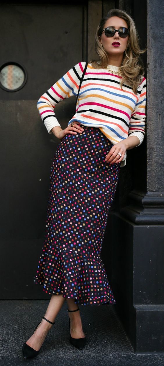 a stylish woman stands against a dark background, showcasing a vibrant outfit. she's wearing a horizontally striped, creamcolored sweater with multicolored stripes, paired with a dark midilength skirt featuring a colorful polka dot pattern. the skirt has a slight mermaid or fishtail hemline. her accessories include black sunglasses, a gold necklace, and delicate rings, complementing her look. she's wearing black anklestrap heels. the overall style exudes confidence and a trendy autumnal aesthetic. the image is welllit, highlighting the textures and colors of the clothing.