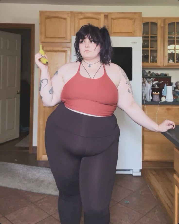 a plussize woman stands indoors in what appears to be a kitchen. she is wearing a red halter top and dark leggings. she has tattoos on both arms and her hair is styled in pigtails. she is holding a banana in one hand. the kitchen features wooden cabinets, a white refrigerator, a countertop, and tiled flooring. a doorway is visible in the background.