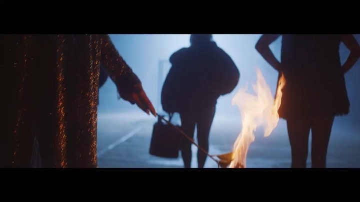 the image shows three figures standing in what appears to be a street, illuminated by a fire. one person is partially visible on the left, wearing a shiny garment, and another is holding a bag. the figures are silhouetted against a smoky backdrop, and the fire is burning in front of them. the overall scene has a cinematic or dramatic feel.