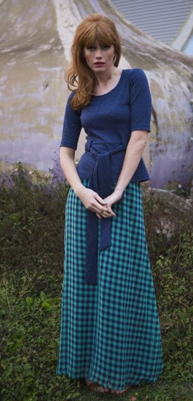 a woman stands in front of a blue and white patterned background. she's wearing a blue blouse and a green and blue plaid skirt, and her hair is in a classic red bob. she is smiling at the camera.