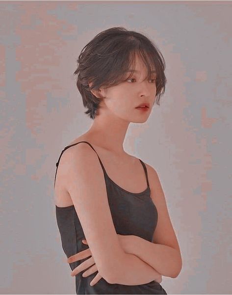 a young woman with short, dark hair styled with a fringe, poses against a textured, muted pink background. she wears a simple black tank top and her arms are crossed, her expression neutral. the lighting is soft and diffused, creating a gentle and feminine atmosphere.