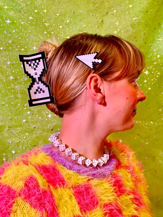 a blonde woman with a side part is wearing a fuzzy pink and yellow sweater. she has an hourglass hair clip and a mouse arrow hair clip in her hair. she is also wearing a dice necklace. the background is a blurred green with white specks. the photo was taken from the side so the woman's profile is showing.