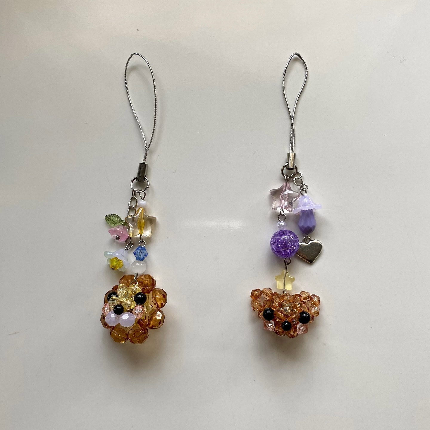two beaded phone charms are displayed on a white surface. the charms feature beaded animals, one appears to be a full bear face and the other resembles a dog. each charm has a silver strap and is decorated with additional beaded details, including flowers, stars, and hearts. the beads used in the charms are various colors, including brown, purple, white, and pink.