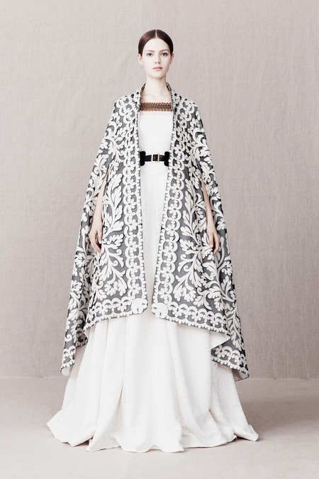 a woman in a white gown with a black lace cape is posing in a studio. she is looking directly at the camera with a serious expression. the cape features floral embroidery. the dress is very simple, and the cape is the main focus of the image. the model is standing on a light gray background. the overall style of the image is elegant and stylish.  the woman is wearing a simple white dress with a belt. the dress is long and flows to the floor. the cape is long and has a lot of detail. the woman's hair is long and dark, and her makeup is simple. the woman is looking directly at the camera. the lighting in the image is soft and diffused, creating a romantic mood.