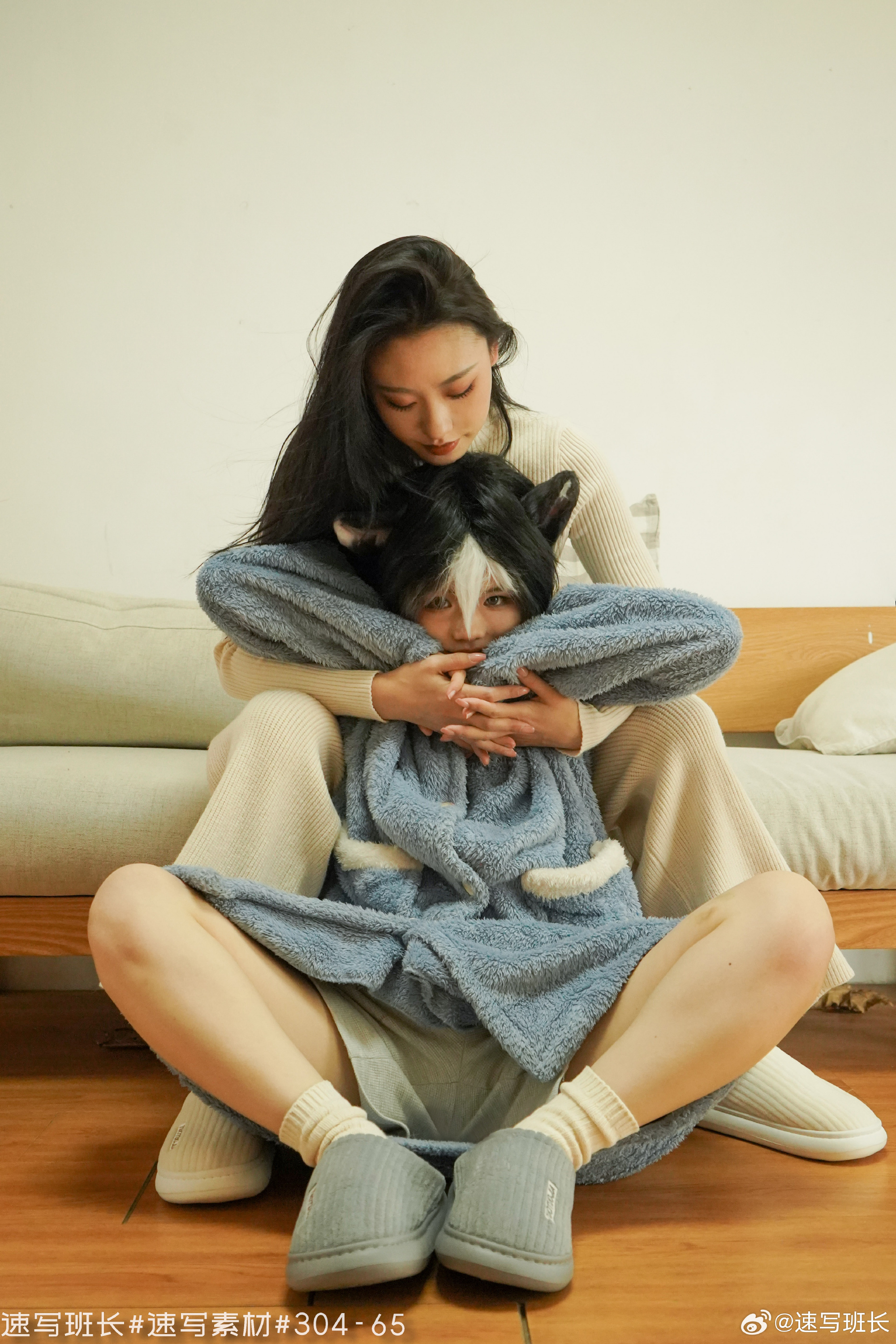 two women are sitting on a wooden floor, one hugging the other from behind. they are both wearing comfortable loungewear, one in white and the other in light blue. the woman in front is wearing a funny animal hat, adding a playful touch to the scene. the atmosphere is warm and intimate, suggesting a close bond between them. the soft, natural light enhances the sense of comfort and relaxation.