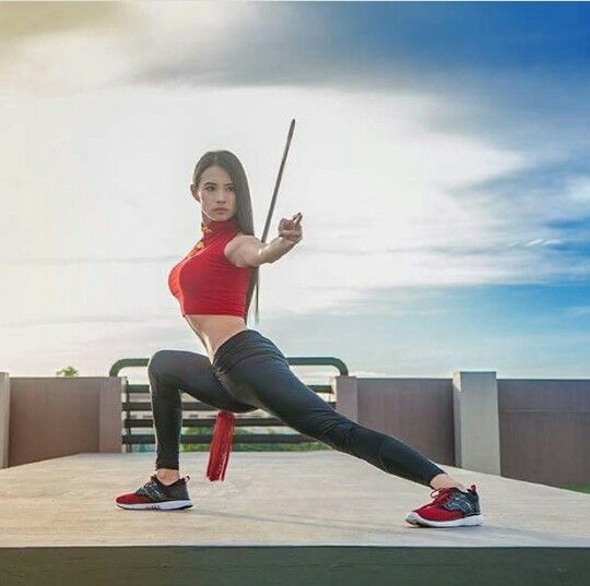 a woman in a red crop top and black leggings performs a martial arts move with a wooden staff, on a rooftop with a blue sky and white clouds in the background. she is  confident and strong.