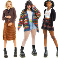 three women are posing in different outfits, all featuring cardigans and skirts. the woman on the left is wearing a brown cardigan, an orange and brown plaid skirt, and black boots. the woman in the middle is wearing a colorful striped cardigan, denim shorts, white tights, and black platform boots. the woman on the right is wearing a black cardigan, a red and green plaid skirt, and black boots. they are all styled with a grunge or alternative aesthetic.