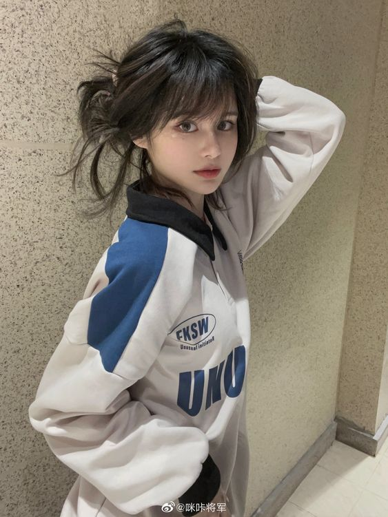 a young woman poses for a selfie in a welllit indoor environment. she has short, dark hair with bangs and wears a white and blue longsleeved shirt with lettering on it. she is looking directly at the camera with a neutral expression.  the image is taken from an eyelevel perspective.