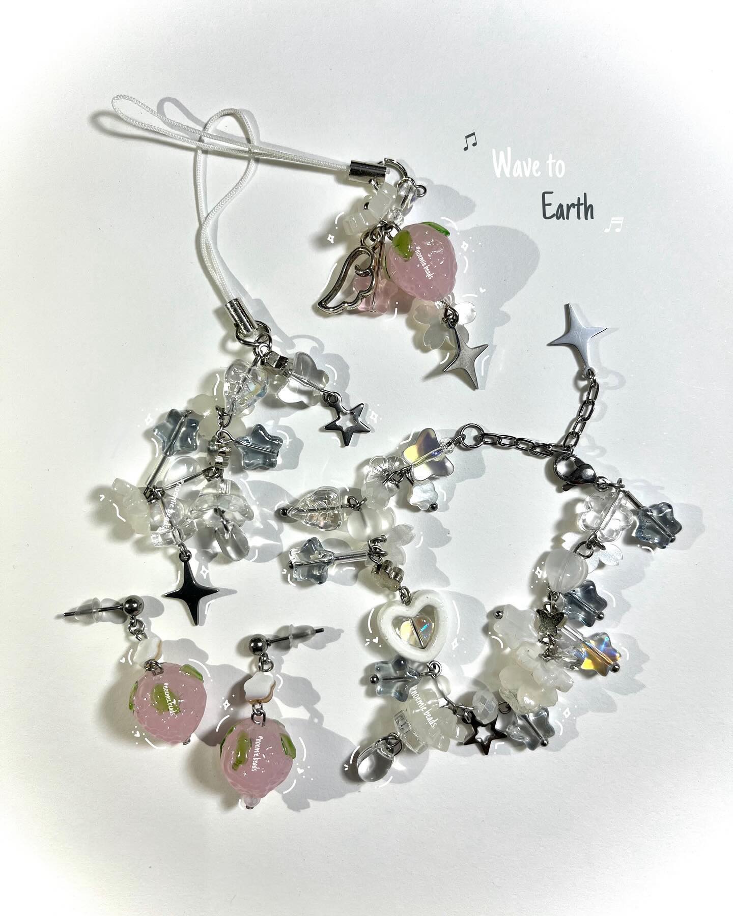 the image shows a collection of handmade jewelry and accessories. there are two pairs of earrings with pink strawberryshaped beads, clear and silver beads, and small black star charms. a charm bracelet features a variety of beads in white, clear, and silver, with star and heart charms. a phone strap is attached to a pink strawberry charm adorned with angel wings and a silver star. the text "wave to earth" and musical notes are placed in the image.