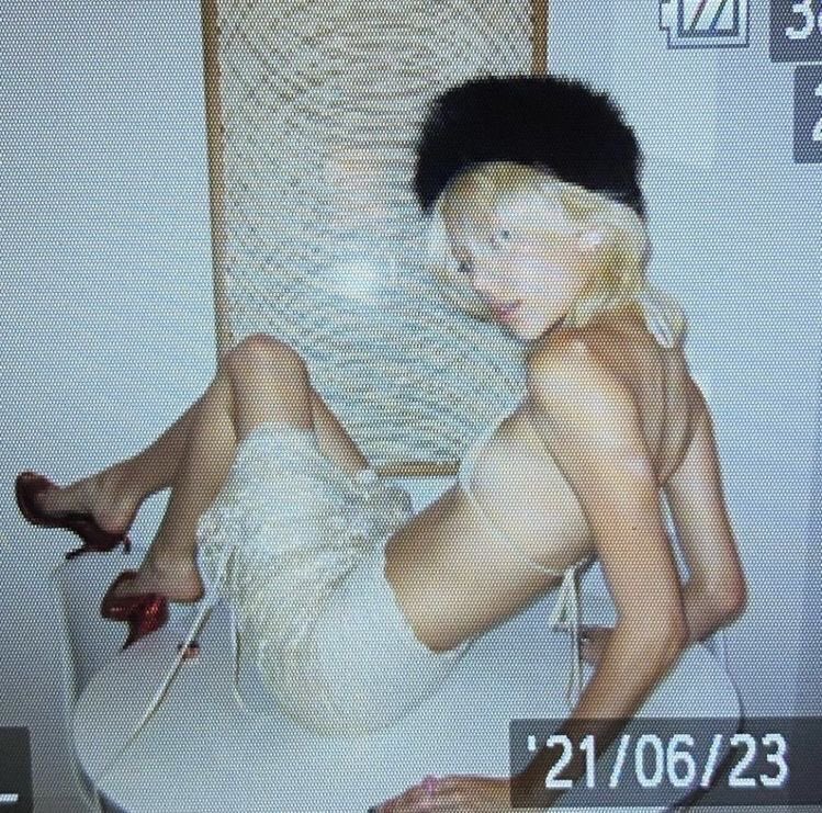 the image is a seemingly scanned photograph or a digital reproduction of a vintage photograph, showing a young blonde woman. she is wearing a black beret, a white slip dress, and red high heels.  the woman is seated, legs slightly crossed, and poses for the camera. the background features a woven, textured wall hanging or screen. the image has a grainy quality consistent with older photographs or film. a date stamp "21/06/23" is visible in the lower right corner, likely indicating when the image was scanned or processed. the overall style is reminiscent of vintage fashion photography, possibly from the mid20th century.